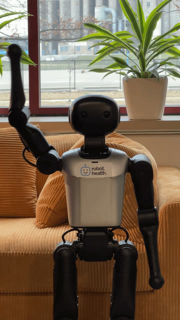 Humanoid robot waving with one arm raised in front of a couch and window