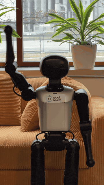 Humanoid robot waving with one arm raised in front of a couch and window